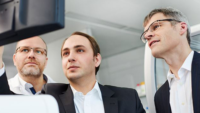 Three men &mdash; including two co-founders of Noscendo, Dr.  Philip Stevens and Dr. Peter Haug &mdash; look at a computer screen together. 