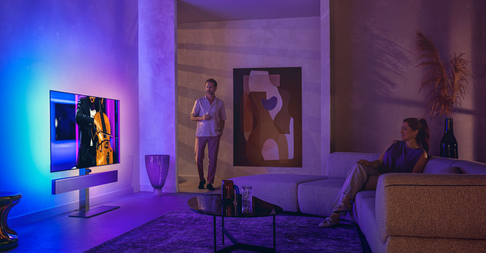 Ukrainian: Чоловік і жінка у стильній вітальні дивляться телевізор Philips Ambilight, на екрані якого виконується композиція на віолончелі.