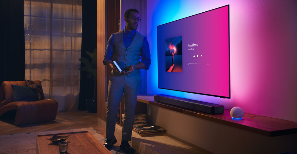 Людина в сучасній вітальні поруч із телевізором Philips Ambilight з музичним інтерфейсом і звуковою панеллю Fidelio.