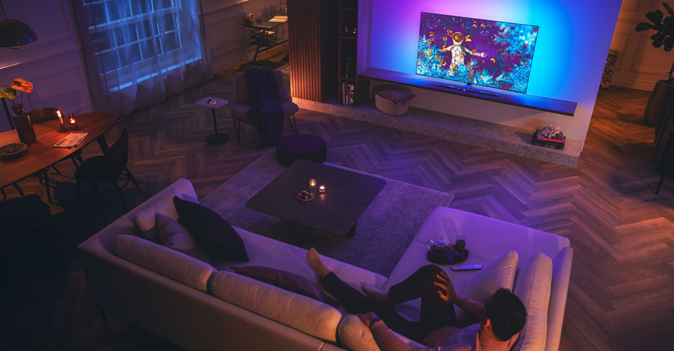 Чоловік розслабляється на дивані у слабо освітленій вітальні, дивлячись телевізор Philips Ambilight.