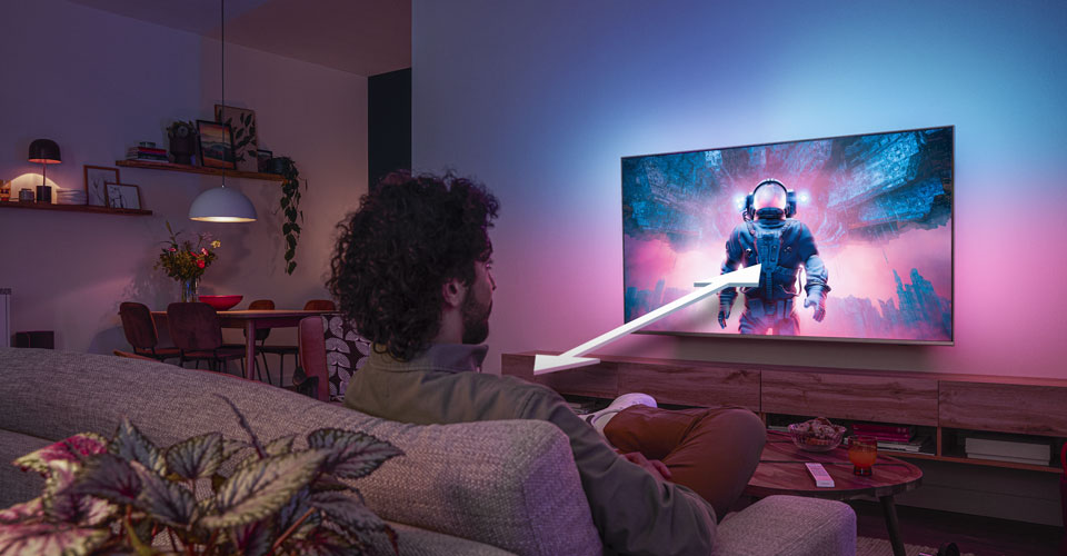 Чоловік сидить на дивані й дивиться телевізор Philips Ambilight, на екрані якого яскрава сцена з астронавтом, зі стрілкою, що вказує оптимальну відстань перегляду.